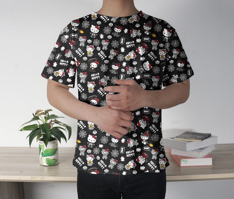 May include: Black t-shirt featuring a repeating pattern of Hello Kitty characters in various Halloween-themed costumes. The design includes spiderwebs, bats, and the words "Hello Kitty" in white. The shirt has short sleeves and a crew neck.