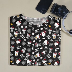 May include: Black t-shirt featuring a Halloween-themed pattern with Hello Kitty characters dressed as mummies, witches, and other spooky figures. The design includes spiderwebs, bats, and the text "Hello Kitty".