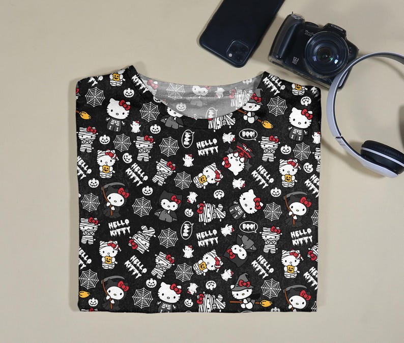 May include: Black t-shirt featuring a Halloween-themed pattern with Hello Kitty characters dressed as mummies, witches, and other spooky figures. The design includes spiderwebs, bats, and the text "Hello Kitty".