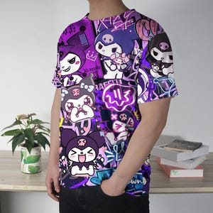 May include: A purple t-shirt with a vibrant graphic design of cartoon characters and graffiti-style text. The shirt has a crew neck and short sleeves. The design features characters in purple, white, and black, creating a bold visual statement.