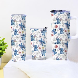 May include: Three white tumblers featuring a repeating pattern of blue cartoon character Stitch, flowers, and leaves. The tumblers vary in size, including a tall, slim tumbler, a shorter tumbler with a lid, and a large mug with a handle.