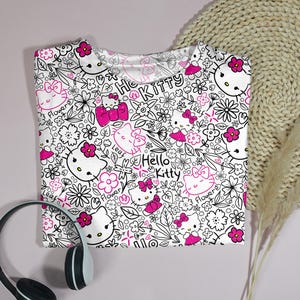 Op de afbeelding: Wit T-shirt met een herhaald patroon van Hello Kitty-personages, bloemen en strikken in zwart en roze. De woorden "Hello Kitty" zijn ook in het ontwerp verwerkt. Een zwart-witte koptelefoon staat op de voorgrond.
