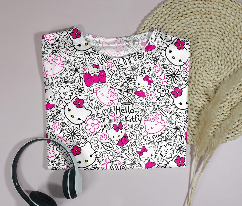 Op de afbeelding: Wit T-shirt met een herhaald patroon van Hello Kitty-personages, bloemen en strikken in zwart en roze. De woorden "Hello Kitty" zijn ook in het ontwerp verwerkt. Een zwart-witte koptelefoon staat op de voorgrond.