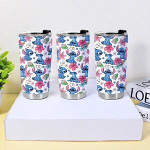 Puede incluir: Tres vasos de viaje de acero inoxidable con tapas negras. Cada vaso presenta un fondo blanco con un patr&oacute;n repetido de personajes de dibujos animados azules, flores rosas y hojas verdes.