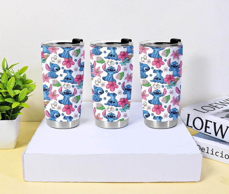 Puede incluir: Tres vasos de viaje de acero inoxidable con tapas negras. Cada vaso presenta un fondo blanco con un patr&oacute;n repetido de personajes de dibujos animados azules, flores rosas y hojas verdes.