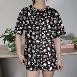 May include: Black t-shirt featuring a repeating pattern of Hello Kitty characters in various poses, with Halloween-themed elements. The design includes white, red, and yellow accents on a black background. The shirt has short sleeves and a crew neck.