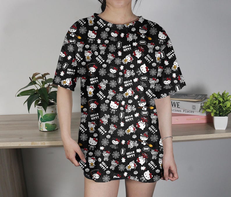 May include: Black t-shirt featuring a repeating pattern of Hello Kitty characters in various poses, with Halloween-themed elements. The design includes white, red, and yellow accents on a black background. The shirt has short sleeves and a crew neck.