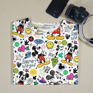 May include: White t-shirt featuring a colorful Mickey Mouse design with various cartoon elements. The design includes Mickey Mouse figures, smiley faces, stars, hearts, and text such as "here we go!" and "Mickey Mouse".