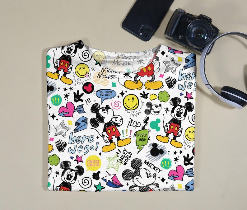May include: White t-shirt featuring a colorful Mickey Mouse design with various cartoon elements. The design includes Mickey Mouse figures, smiley faces, stars, hearts, and text such as "here we go!" and "Mickey Mouse".