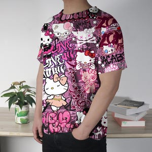 May include: A short-sleeved t-shirt with a vibrant, all-over print featuring Hello Kitty characters in various poses, set against a pink and purple graffiti-style background. The design includes text elements and other graphic details.