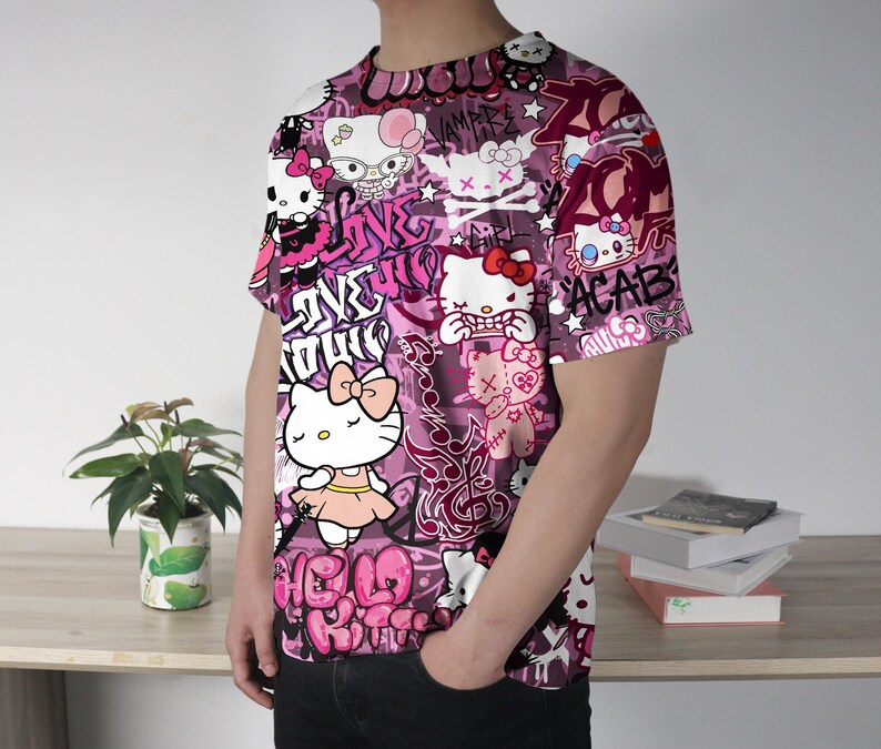 May include: A short-sleeved t-shirt with a vibrant, all-over print featuring Hello Kitty characters in various poses, set against a pink and purple graffiti-style background. The design includes text elements and other graphic details.