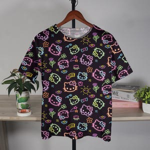 May include: Black t-shirt with a repeating neon-style pattern of Hello Kitty faces, flowers, suns, and bows. The design features bright pink, yellow, green, and blue accents. The shirt is hanging on a wooden hanger.