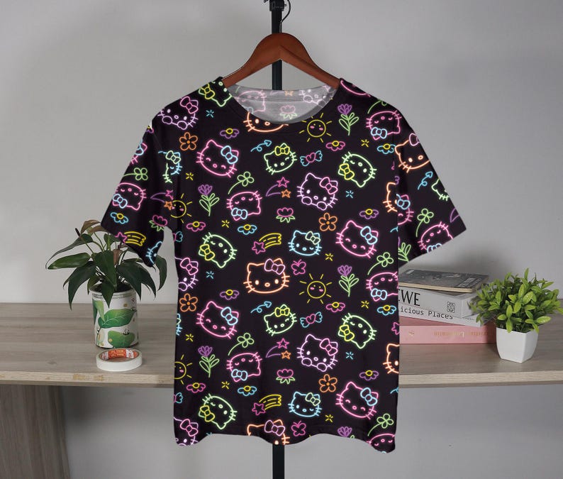 May include: Black t-shirt with a repeating neon-style pattern of Hello Kitty faces, flowers, suns, and bows. The design features bright pink, yellow, green, and blue accents. The shirt is hanging on a wooden hanger.