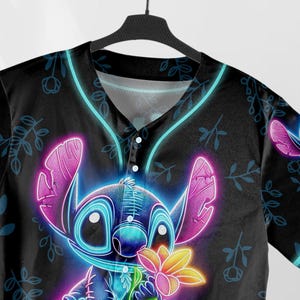 Puede incluir: Una camiseta de b&eacute;isbol negra con un personaje Stitch de color azul ne&oacute;n y rosa sosteniendo una flor. El fondo es negro con un patr&oacute;n floral.