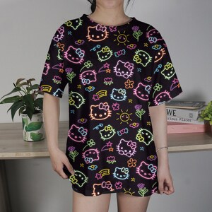 May include: Black oversized t-shirt featuring a neon-style pattern of Hello Kitty faces, flowers, stars, and suns. The design includes pink, yellow, green, and orange accents. The shirt has short sleeves and a round neckline.