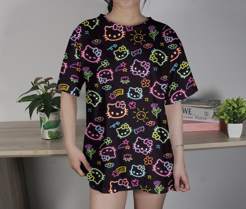 May include: Black oversized t-shirt featuring a neon-style pattern of Hello Kitty faces, flowers, stars, and suns. The design includes pink, yellow, green, and orange accents. The shirt has short sleeves and a round neckline.