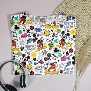 May include: White crew neck t-shirt featuring a colorful Mickey Mouse pattern. The design includes Mickey Mouse illustrations, smiley faces, hearts, stars, and the text "Here we go!" and "Mickey Mouse". Black and white headphones are in the foreground.