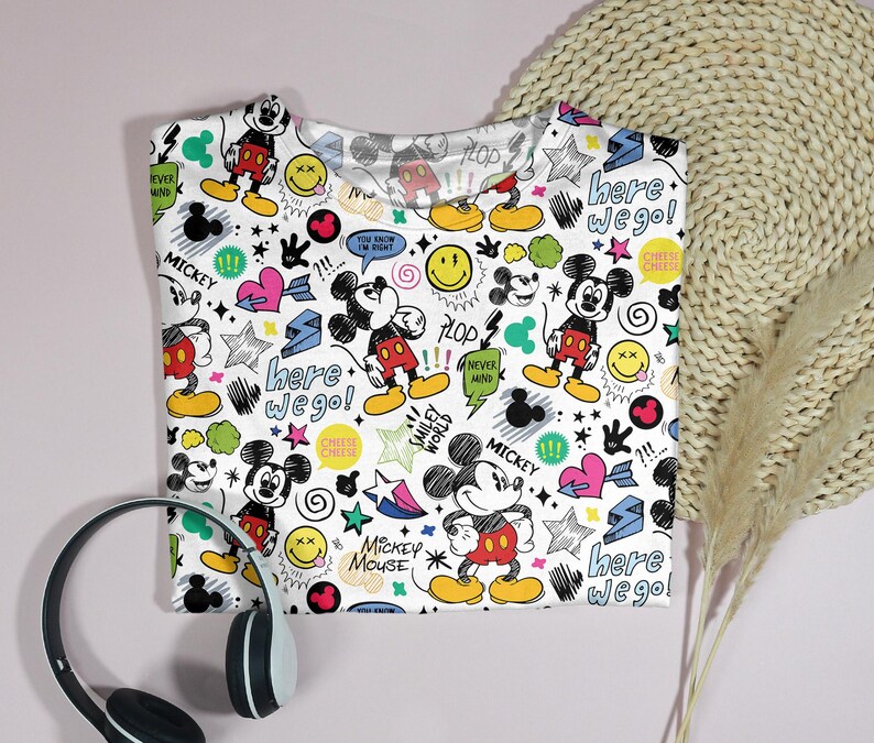 May include: White crew neck t-shirt featuring a colorful Mickey Mouse pattern. The design includes Mickey Mouse illustrations, smiley faces, hearts, stars, and the text "Here we go!" and "Mickey Mouse". Black and white headphones are in the foreground.