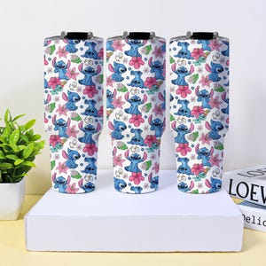 Puede incluir: Tres vasos aislados con fondo blanco, con un patr&oacute;n repetido de personajes de dibujos animados azules, flores rosas y hojas verdes. Cada vaso tiene una tapa negra.