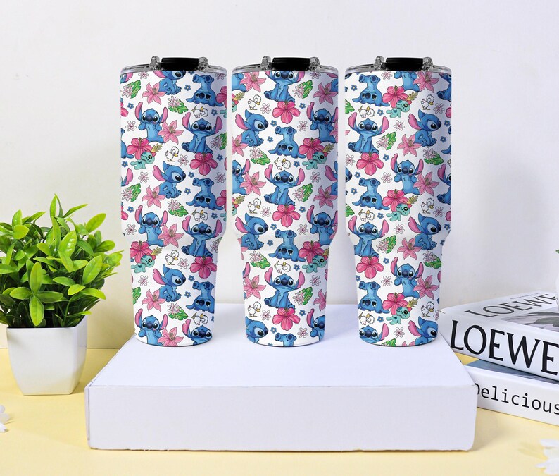 Puede incluir: Tres vasos aislados con fondo blanco, con un patr&oacute;n repetido de personajes de dibujos animados azules, flores rosas y hojas verdes. Cada vaso tiene una tapa negra.
