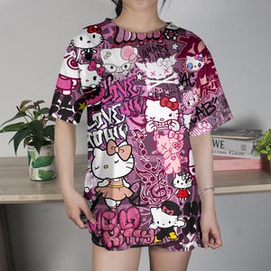 May include: A short-sleeved, oversized t-shirt featuring a vibrant, all-over print of Hello Kitty characters in various poses, set against a backdrop of graffiti-style text and designs in shades of pink, purple, and white. The shirt has a crew neck.