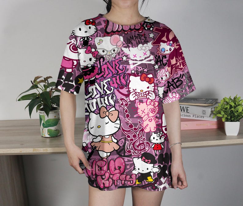 May include: A short-sleeved, oversized t-shirt featuring a vibrant, all-over print of Hello Kitty characters in various poses, set against a backdrop of graffiti-style text and designs in shades of pink, purple, and white. The shirt has a crew neck.