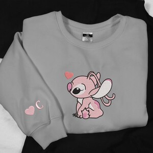 May include: Gray sweatshirt with a pink Stitch character wearing a pink heart. The sweatshirt has a pink heart and the letter C embroidered on the sleeve.