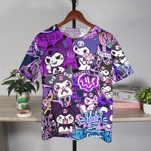 May include: A purple and white graphic t-shirt with cartoon characters and graffiti art. The t-shirt has a crew neck and short sleeves. The design includes the word "Crown" in blue and other text.