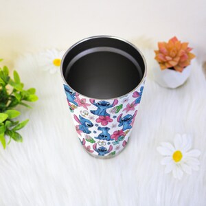 Puede incluir: Un vaso de acero inoxidable con un patr&oacute;n repetido del personaje Stitch de Lilo & Stitch, junto con flores rosas y moradas. El vaso tiene un borde plateado y un interior oscuro.