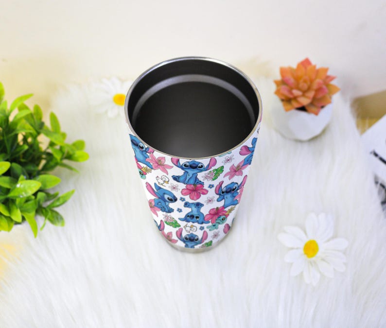 Puede incluir: Un vaso de acero inoxidable con un patr&oacute;n repetido del personaje Stitch de Lilo & Stitch, junto con flores rosas y moradas. El vaso tiene un borde plateado y un interior oscuro.