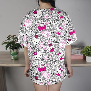 Op de afbeelding: Wit T-shirt met een herhaald patroon van Hello Kitty gezichten, roze strikken, bloemen en de woorden "Hello Kitty" in een zwarte omtrek. Het shirt heeft korte mouwen en een losse pasvorm.