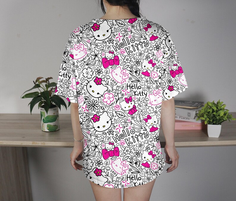 Op de afbeelding: Wit T-shirt met een herhaald patroon van Hello Kitty gezichten, roze strikken, bloemen en de woorden "Hello Kitty" in een zwarte omtrek. Het shirt heeft korte mouwen en een losse pasvorm.