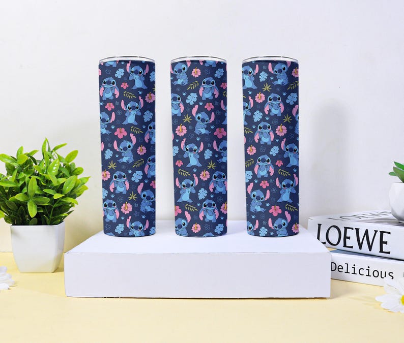 Puede incluir: Tres vasos cil&iacute;ndricos con un fondo azul marino que presenta un patr&oacute;n repetido de un personaje de dibujos animados azul, flores rosas y hojas verdes. Los vasos se exhiben sobre una plataforma blanca.