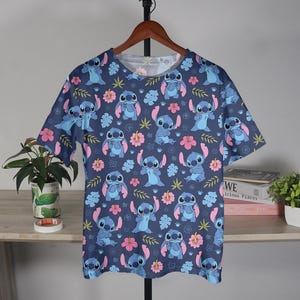 Pu&ograve; includere: Una maglietta blu a maniche corte con un motivo ripetuto del personaggio Stitch di Lilo & Stitch, fiori rosa e foglie verdi. La maglietta &egrave; appesa a una gruccia di legno.