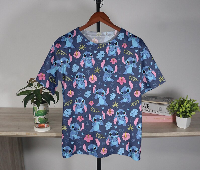 Pu&ograve; includere: Una maglietta blu a maniche corte con un motivo ripetuto del personaggio Stitch di Lilo & Stitch, fiori rosa e foglie verdi. La maglietta &egrave; appesa a una gruccia di legno.
