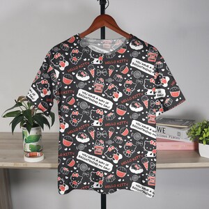 May include: Black t-shirt featuring a repeating pattern of Hello Kitty, cupcakes, ice cream cones, and candies. The shirt includes text bubbles that say "You Have a Way of Brightening My Day!" and "Hello Kitty."
