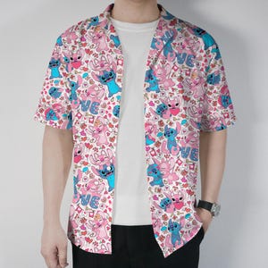 Puede incluir: Una camisa de manga corta con un estampado repetido de personajes de dibujos animados en rosa y azul sobre un fondo blanco. La camisa tiene cuello de campamento y se usa abierta sobre una camiseta blanca.