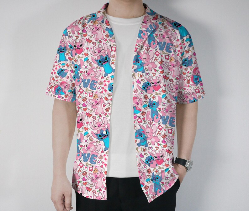 Puede incluir: Una camisa de manga corta con un estampado repetido de personajes de dibujos animados en rosa y azul sobre un fondo blanco. La camisa tiene cuello de campamento y se usa abierta sobre una camiseta blanca.