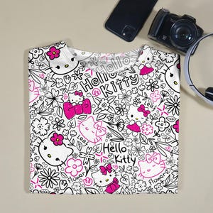 Op de afbeelding: Wit T-shirt met een herhaald patroon van Hello Kitty-personages, bloemen en de woorden "Hello Kitty". Het ontwerp maakt gebruik van zwarte contouren en roze accenten. Het shirt is opgevouwen en wordt op een neutrale ondergrond gepresenteerd.