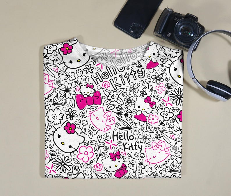 Op de afbeelding: Wit T-shirt met een herhaald patroon van Hello Kitty-personages, bloemen en de woorden "Hello Kitty". Het ontwerp maakt gebruik van zwarte contouren en roze accenten. Het shirt is opgevouwen en wordt op een neutrale ondergrond gepresenteerd.