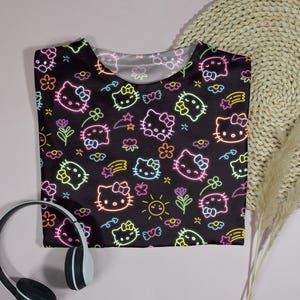 May include: Black t-shirt featuring a neon-style pattern of Hello Kitty faces, flowers, stars, and suns. The design includes pink, yellow, green, and blue accents. A pair of black and white headphones are in the lower left corner.