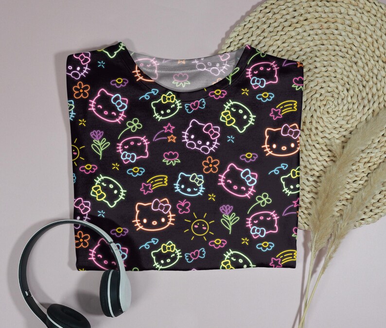 May include: Black t-shirt featuring a neon-style pattern of Hello Kitty faces, flowers, stars, and suns. The design includes pink, yellow, green, and blue accents. A pair of black and white headphones are in the lower left corner.