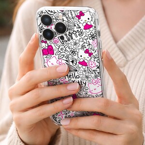 May include: Clear phone case featuring a white and black floral pattern with Hello Kitty characters in pink. The case has the words "Hello Kitty" and "Kitty" in a playful font. The case is held in a person's hands.