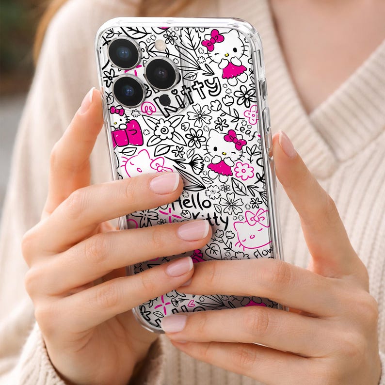 May include: Clear phone case featuring a white and black floral pattern with Hello Kitty characters in pink. The case has the words "Hello Kitty" and "Kitty" in a playful font. The case is held in a person's hands.