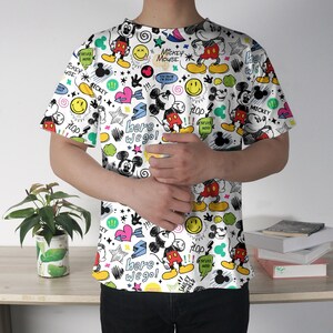 May include: White t-shirt featuring a colorful all-over print of Mickey Mouse, smiley faces, stars, hearts, and text. The text includes "Mickey Mouse", "Here we go!", and "Plop". The shirt has short sleeves and a crew neck.