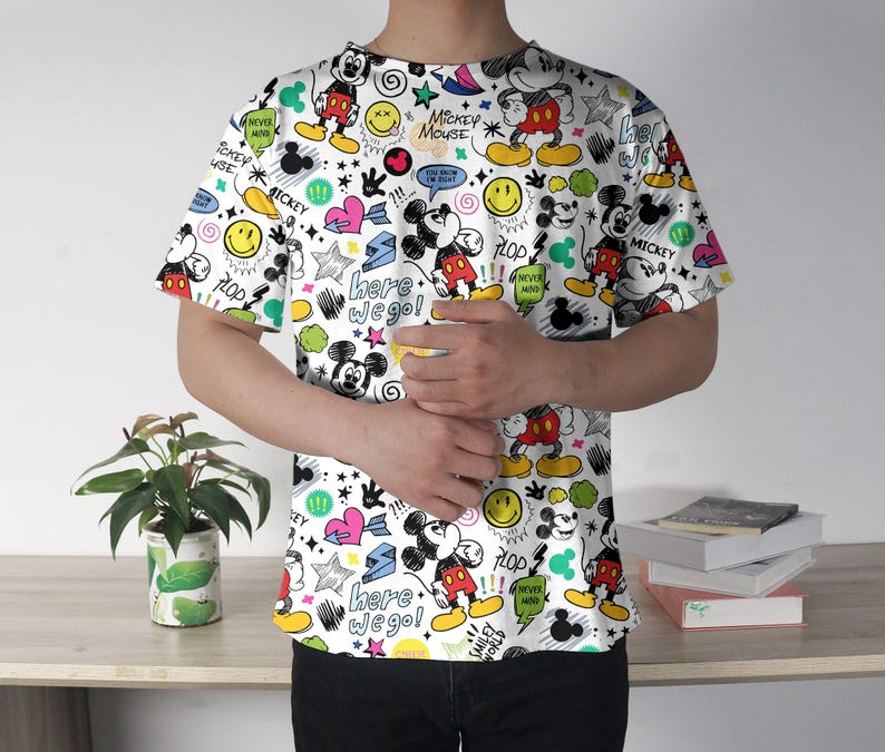 May include: White t-shirt featuring a colorful all-over print of Mickey Mouse, smiley faces, stars, hearts, and text. The text includes "Mickey Mouse", "Here we go!", and "Plop". The shirt has short sleeves and a crew neck.