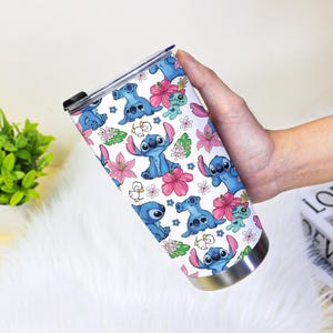 Puede incluir: Un vaso de acero inoxidable con un patr&oacute;n repetido del alien&iacute;gena azul Stitch, flores rosas y hojas verdes sobre un fondo blanco. El vaso tiene una base plateada y una tapa negra.