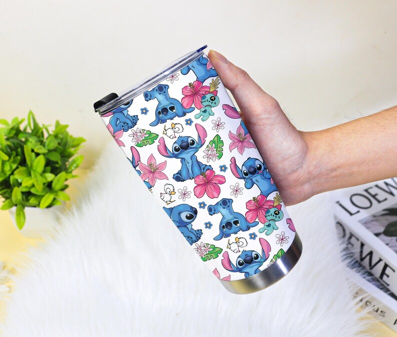Puede incluir: Un vaso de acero inoxidable con un patr&oacute;n repetido del alien&iacute;gena azul Stitch, flores rosas y hojas verdes sobre un fondo blanco. El vaso tiene una base plateada y una tapa negra.