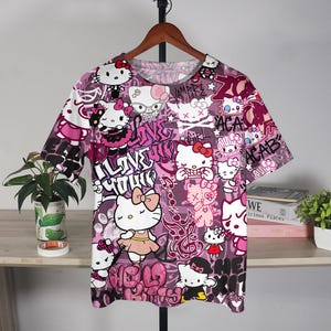 May include: A short-sleeved t-shirt with a vibrant, all-over print featuring the Hello Kitty character in various poses, set against a graffiti-style background with pink and purple hues. The design includes text elements like "I Love You" and "ACAB".