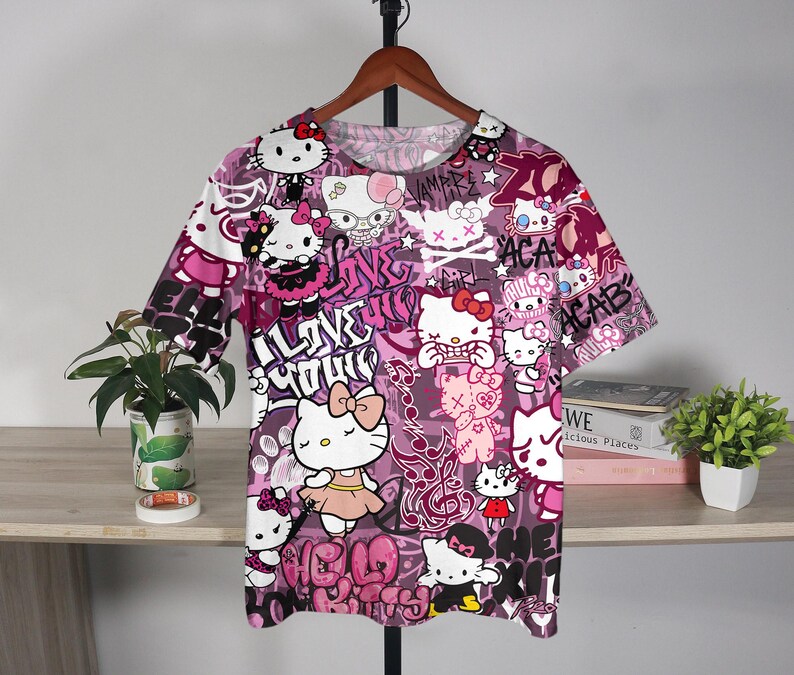 May include: A short-sleeved t-shirt with a vibrant, all-over print featuring the Hello Kitty character in various poses, set against a graffiti-style background with pink and purple hues. The design includes text elements like "I Love You" and "ACAB".